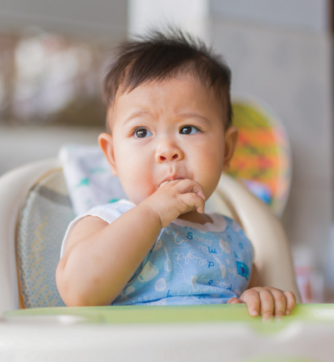 Baby in high chair, wearing a blue bib, sucks thumb thoughtfully.