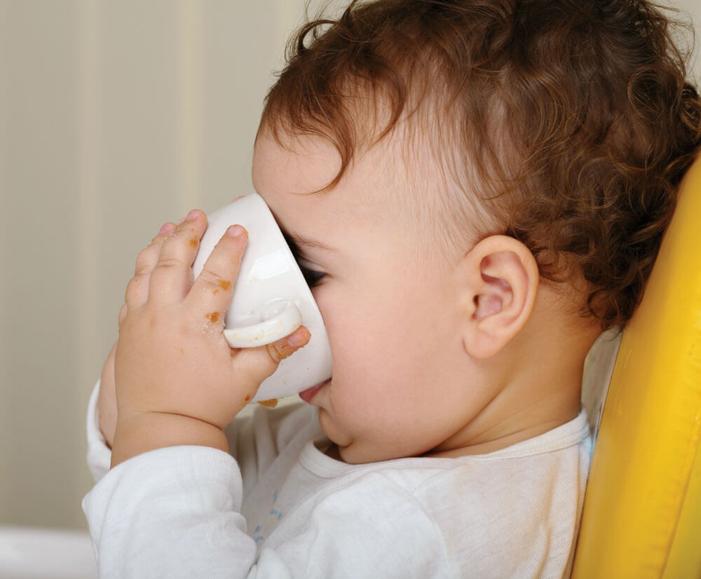 Child drinking from a white cup, seated on a yellow chair, with curls and messy hands.