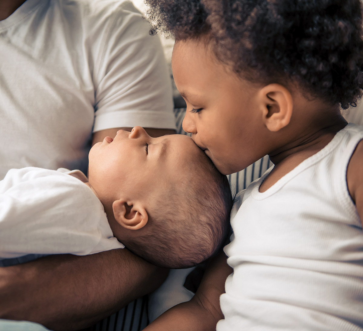 Toddler gently kisses sleeping baby's forehead, held in adult arms.