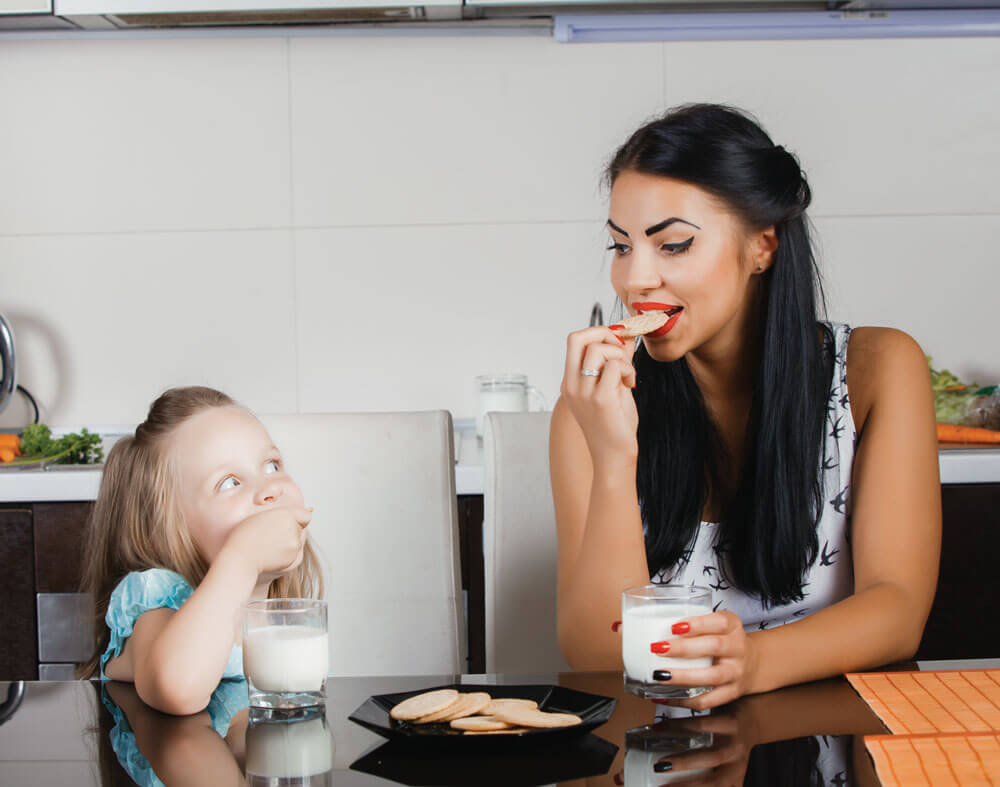 Mother and daughter enjoy milk and cookies together at the kitchen table.