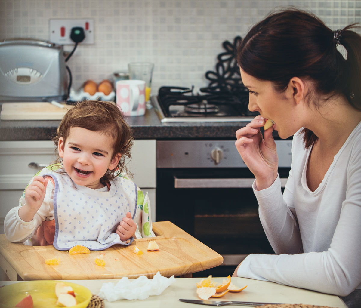 Toddler smiling in high chair while parent eats fruit in kitchen.