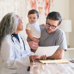 BabyDadVisitDr Doctor discussing paperwork with dad holding a baby in a clinic office.